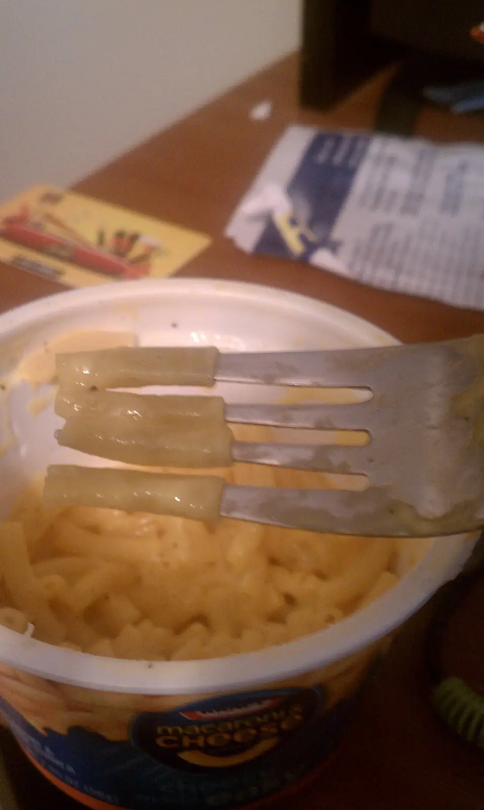 A close-up photo shows a fork lifting three macaroni noodles from a partially eaten cup of mac and cheese. The noodles are…
