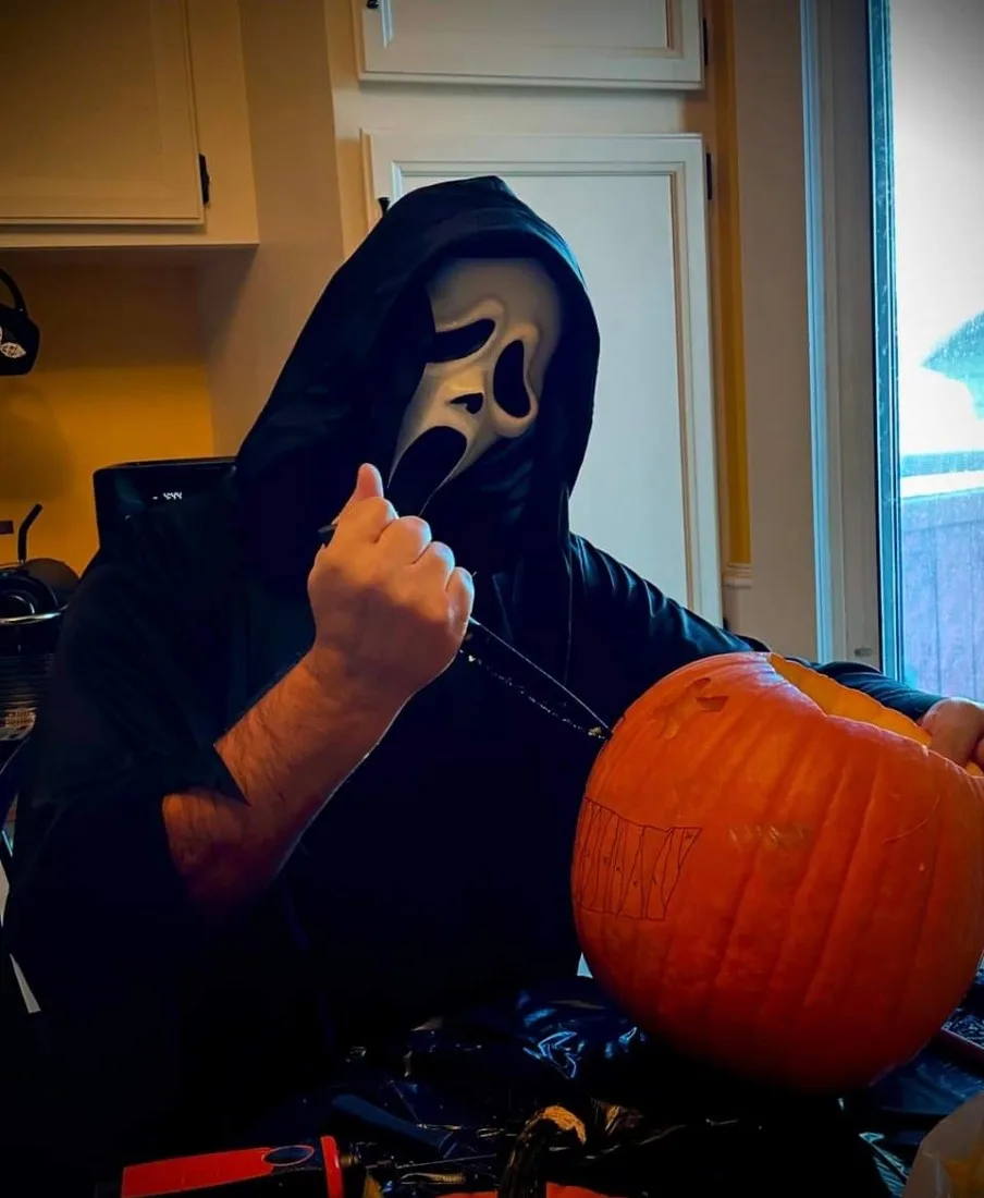 A person wearing a black hooded robe and a white Ghostface mask from the Scream movie franchise is carving a pumpkin in a…