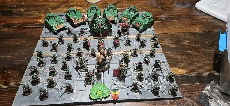 A tabletop wargame setup featuring a large army of green-painted miniature figures, including tanks and infantry, arranged…