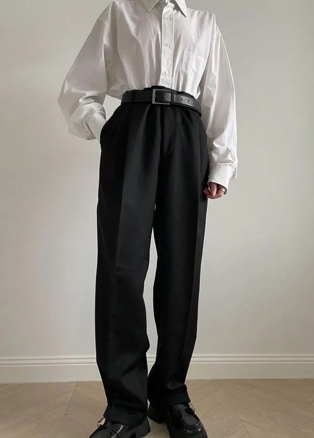 A person wearing a white button-up shirt, black high-waisted trousers, and black loafers stands with hands in pockets. A…