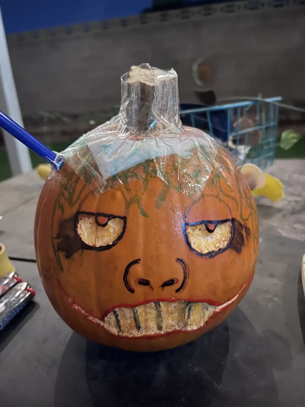 A close-up of an orange pumpkin with a hand-drawn angry face, featuring narrowed eyes, a scowling mouth with teeth, and…