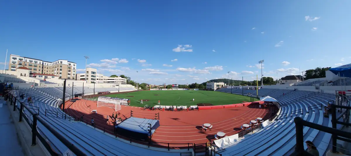 A wide-angle photo of an empty outdoor stadium with a green baseball field, red running track, and white bleachers under a…