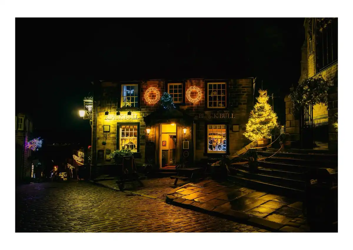 A warmly lit stone pub named 'Black Bull' at night, decorated with glowing Christmas wreaths and a large tree, with…