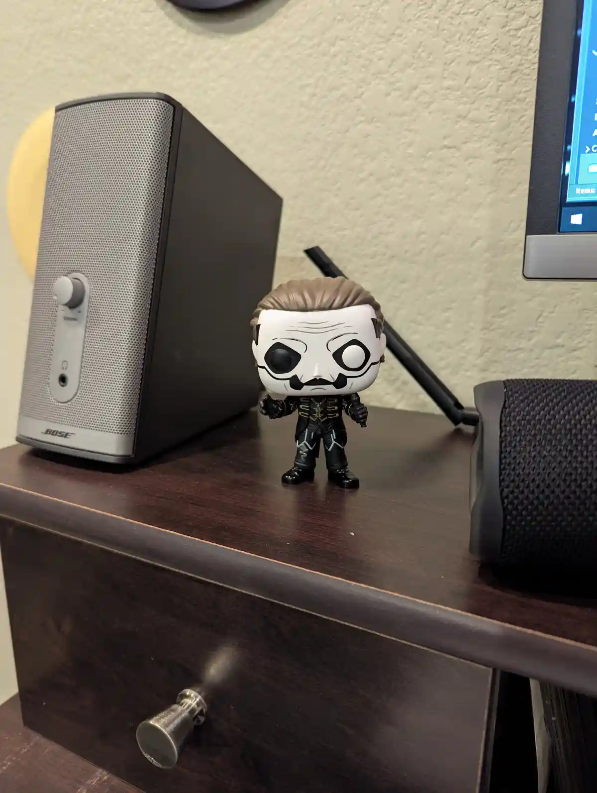 A Funko Pop! figure of the character Ghostface from Scream stands on a dark wooden desk next to a Bose speaker and a…