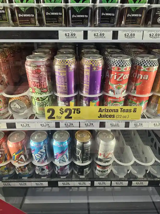 A grocery store beverage aisle displays shelves of Arizona iced teas and juices with a yellow sale sign reading '2 for $2.75…