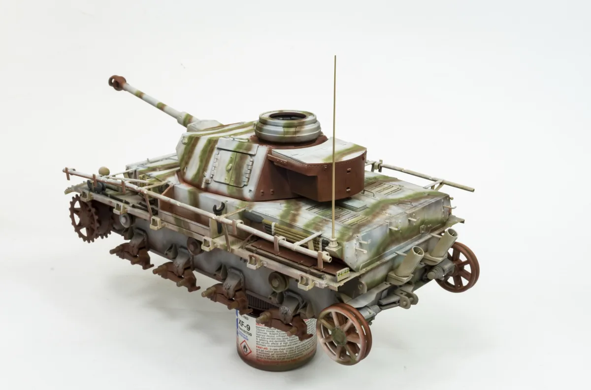 A detailed scale model of a German World War II tank, painted in camouflage, is shown resting on a can of XF-9 paint. The…