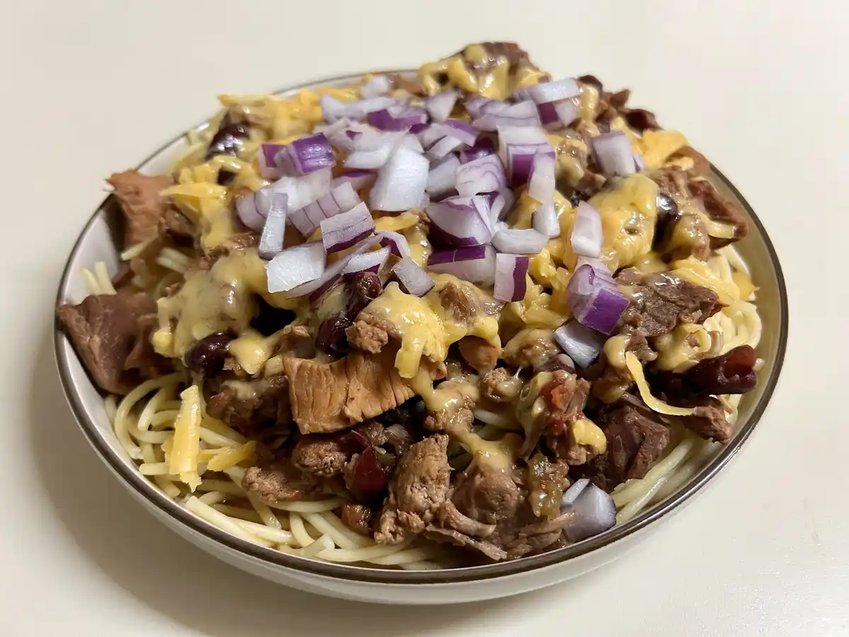 A close-up photo of a bowl filled with spaghetti noodles topped with melted yellow cheese, chunks of cooked meat, dark…
