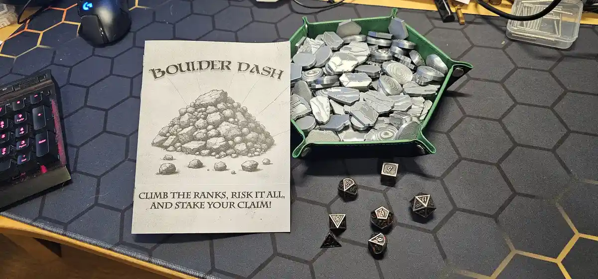 A tabletop scene shows a printed 'Boulder Dash' game box with a pile of rocks illustration and the slogan 'Climb the ranks…