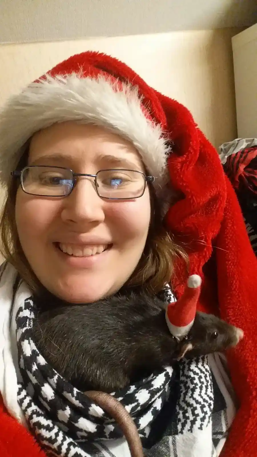 A smiling person wearing glasses and a red Santa hat holds a black rat wrapped in a black-and-white patterned scarf. The rat…