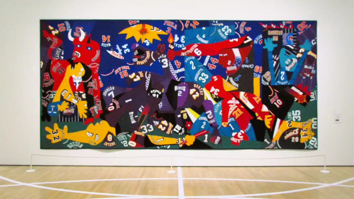 A large quilt displayed in a gallery, made from numerous cut-up basketball jerseys featuring team logos, player names, and…