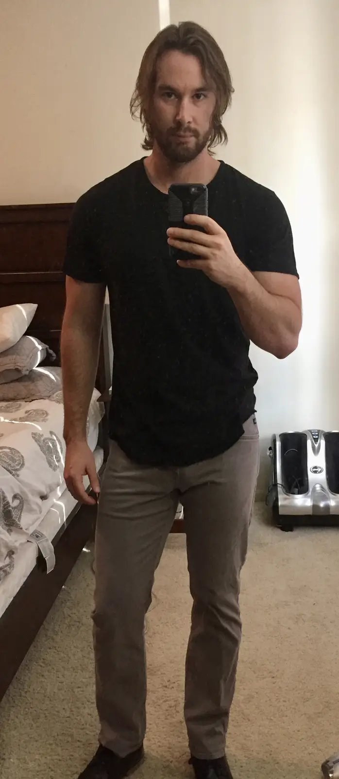 A man with long hair and a beard takes a mirror selfie in a bedroom, wearing a black t-shirt and grey pants, holding a…