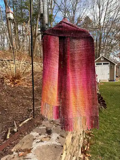 A handwoven, gradient-colored scarf in shades of magenta, pink, and orange hangs outdoors on a stone wall, with a bird…
