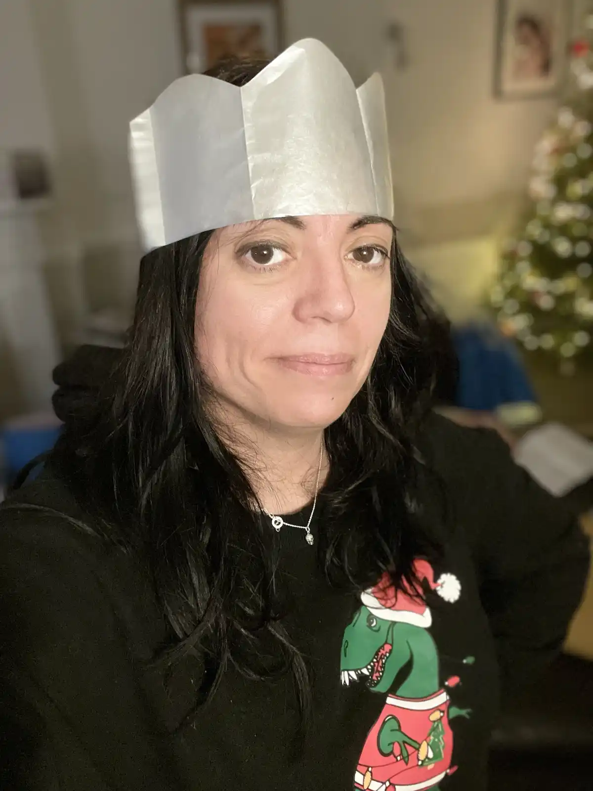 A woman with long dark hair wears a simple, shiny silver paper crown on her head and a black holiday-themed hoodie featuring…