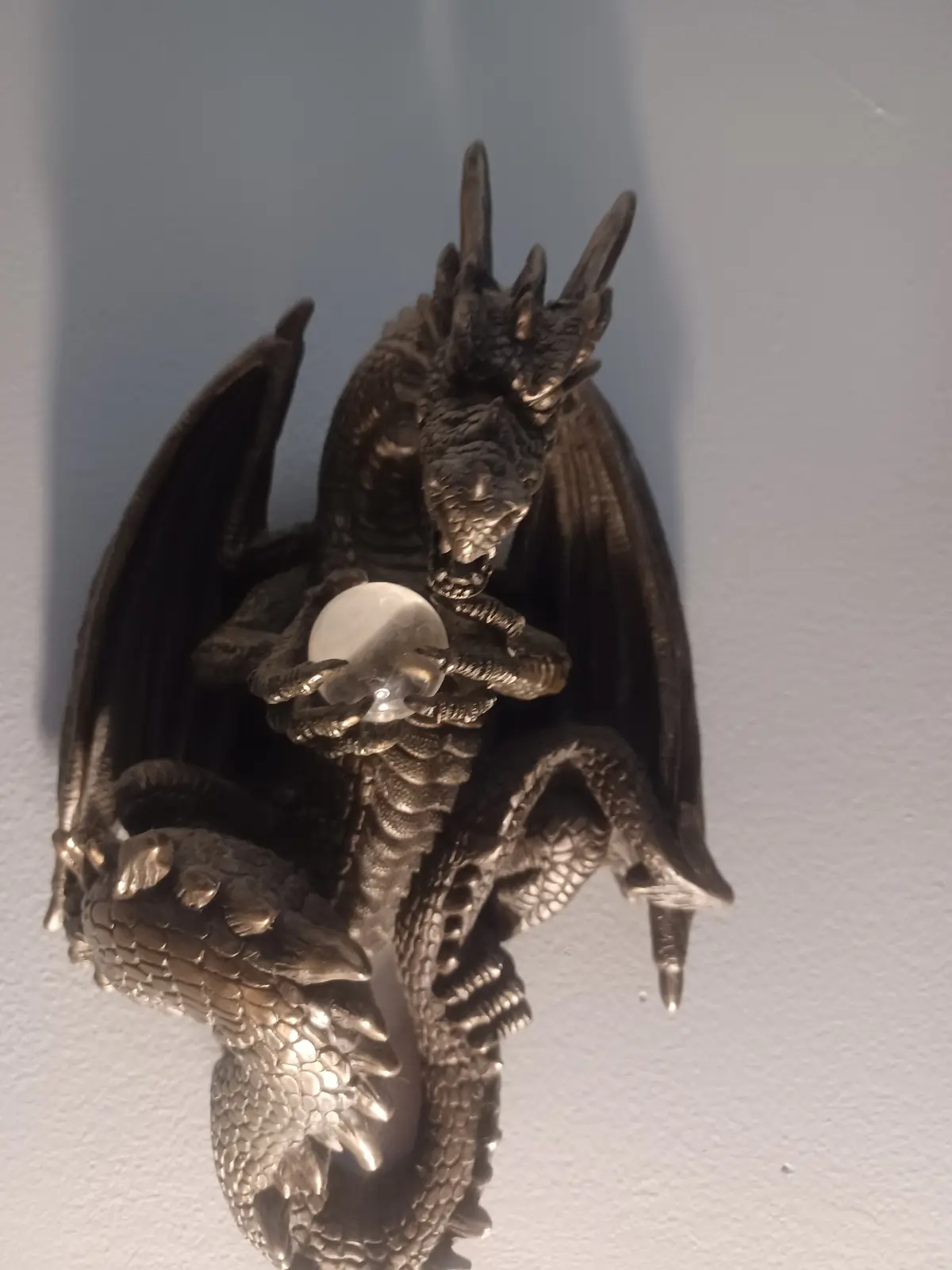 A dark, metallic dragon figurine with wings spread and claws gripping a clear crystal ball, mounted on a plain wall. The…
