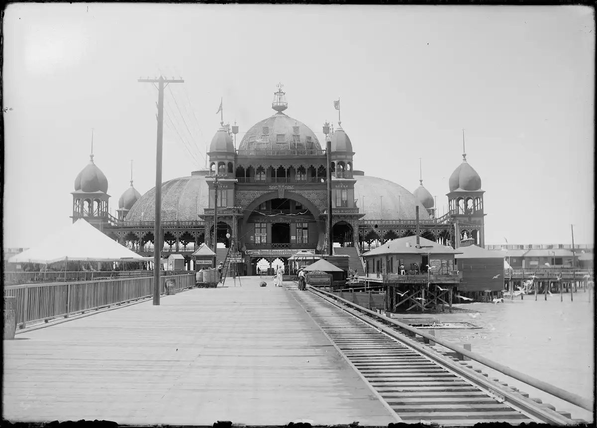 A black-and-white historical photo shows a grand, ornate pier building with multiple domes and flags, viewed from a wooden…