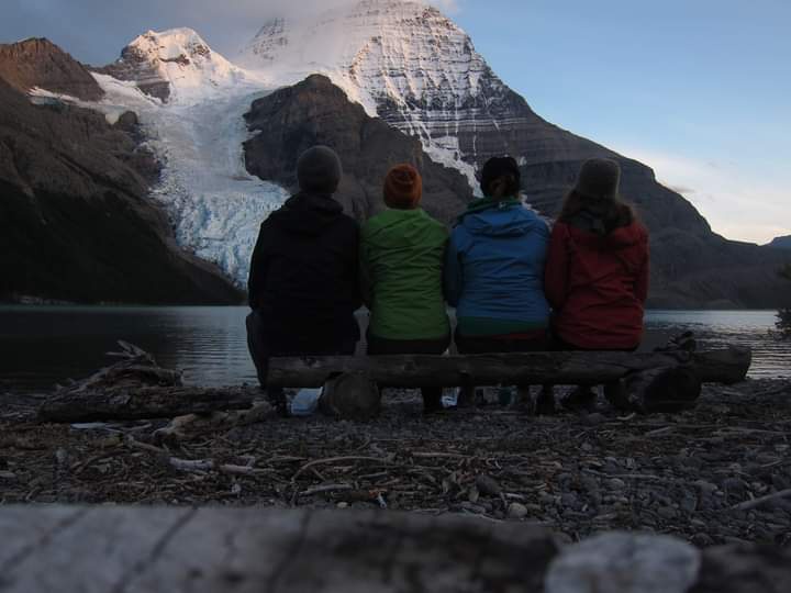 four people sit side-by-side on a log bench facing a massive, snow-capped mountain and glacier. They are dressed in warm…