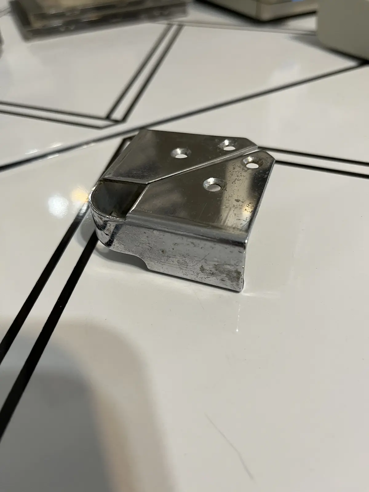 A close-up photo shows a shiny, silver metal corner bracket resting on a white tiled floor with black grout lines. The…