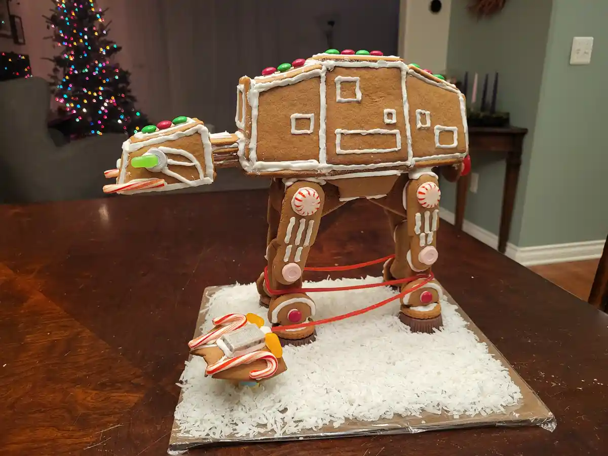A gingerbread model of an AT-AT walker from Star Wars is displayed on a wooden table, decorated with white icing, candy…