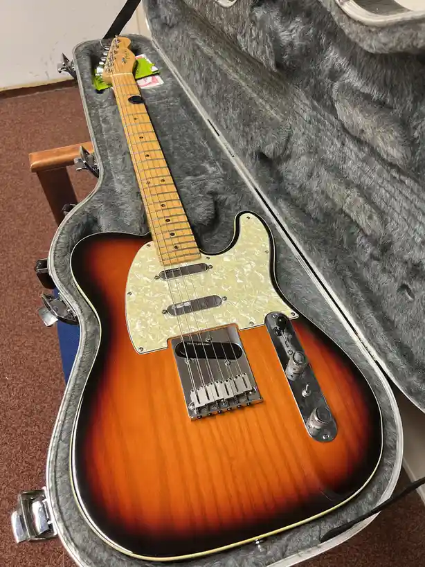 A sunburst-finish electric guitar with a light-colored pickguard and maple neck rests inside an open, grey-felt-lined hard…