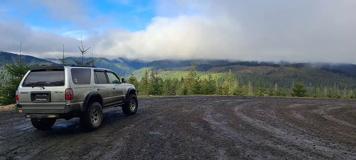 A silver, lifted Toyota 4Runner SUV is parked on a wet, gravel overlook with a scenic view of forested mountains partially…