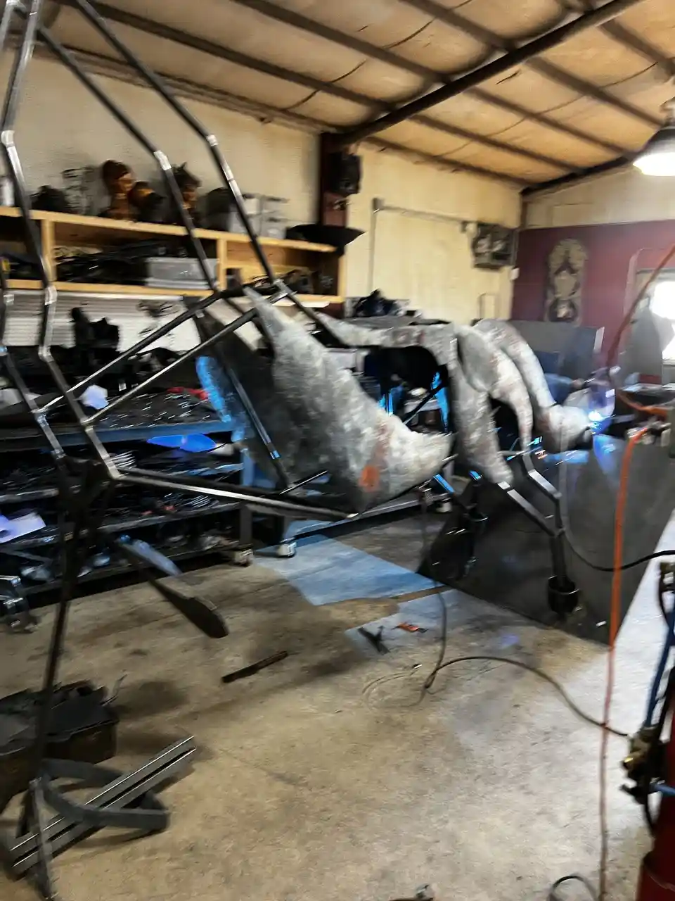 A blurry photo of a cluttered garage workshop with a partially built metal car frame being welded, showing sparks and tools…