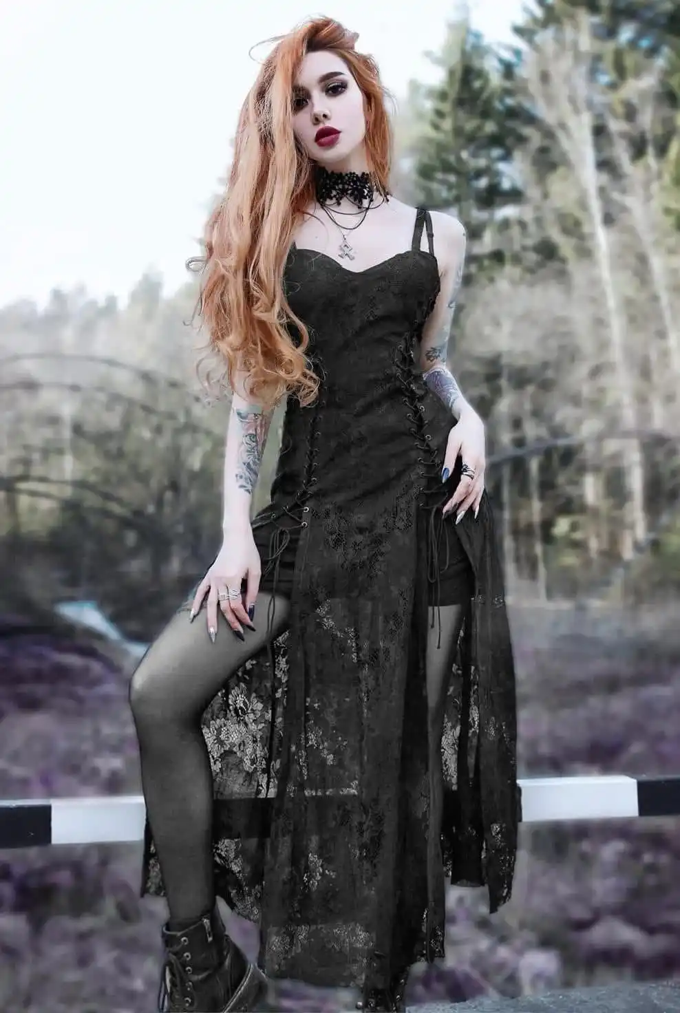 A woman with long orange hair, wearing a black lace dress, choker, and boots, poses outdoors near a railing. She has tattoos…