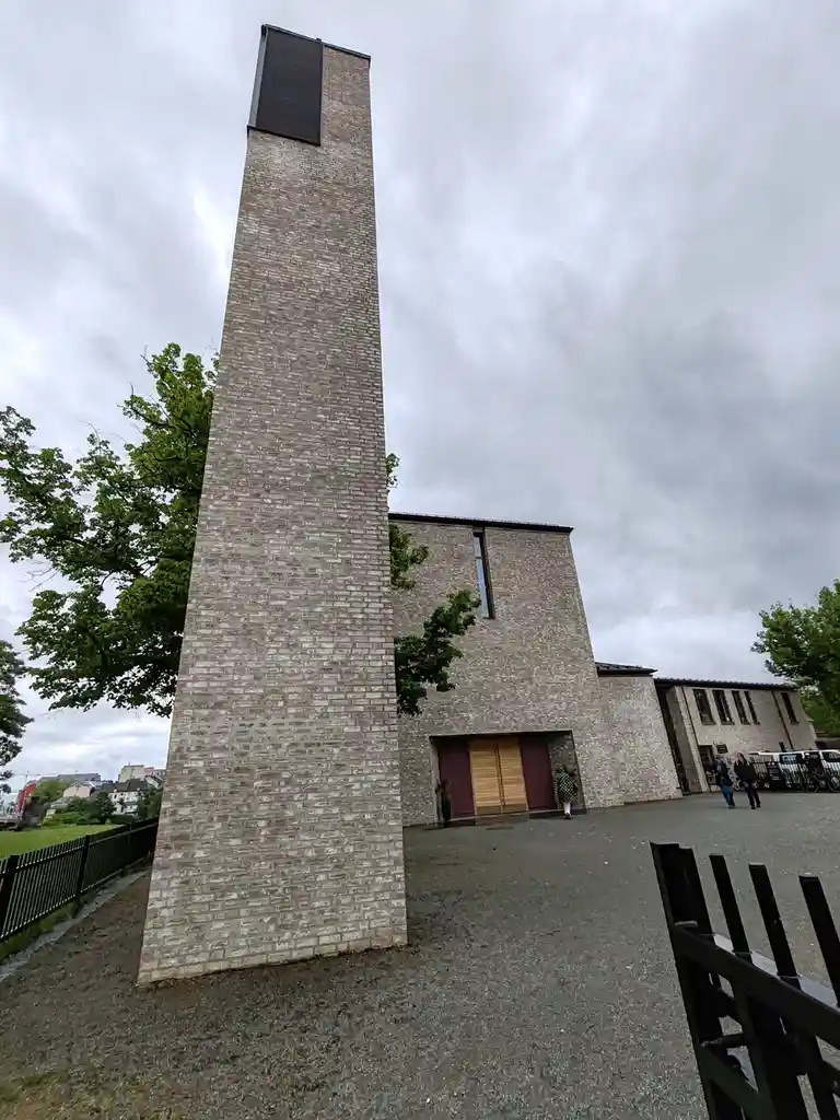 A low-angle photo shows a tall, narrow brick tower attached to a modern brick church building under an overcast sky. The…
