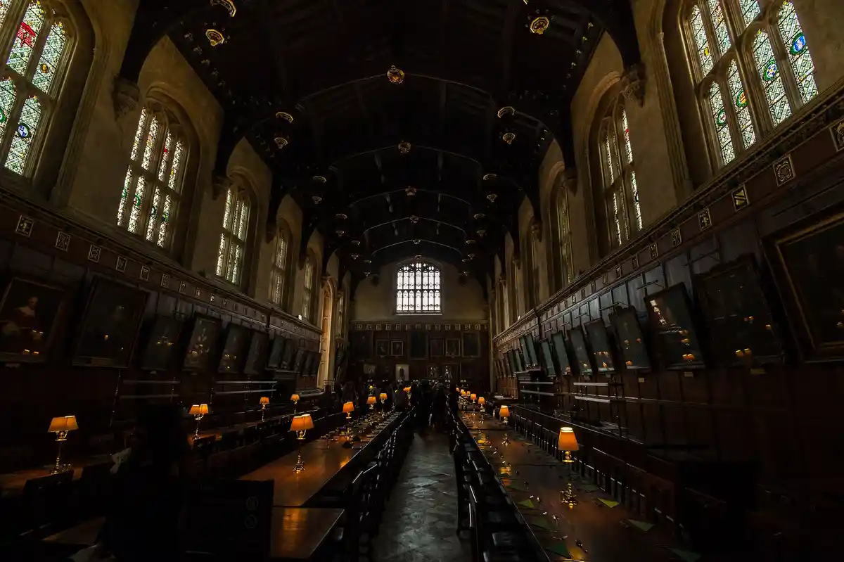 A dimly lit, grand historic dining hall with long wooden tables, ornate stained glass windows, and portraits on the walls…