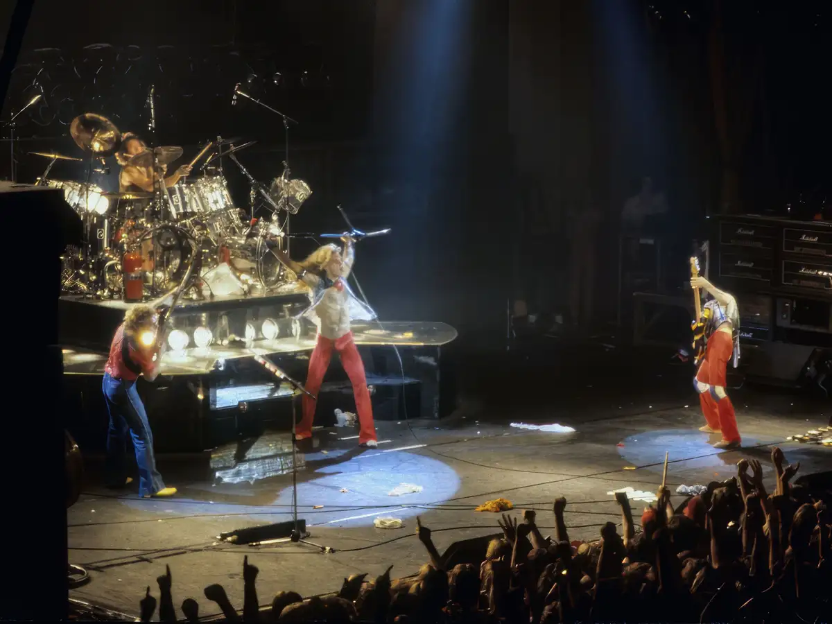 A vintage photo captures a rock band performing on a lit stage with a drummer, a lead singer in red pants, and a guitarist…