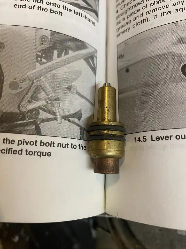 A brass mechanical part with rubber seals sits balanced on the open pages of a repair manual, which contain black-and-white…