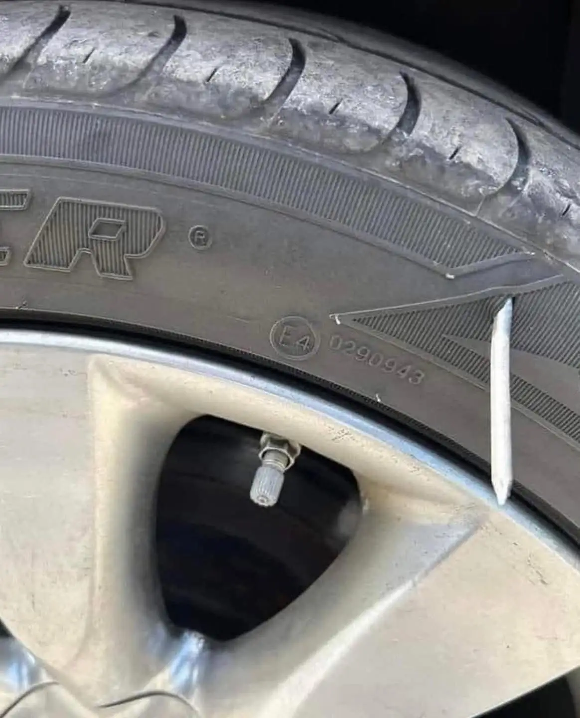 A close-up photo of a car tire with a white nail stuck in its sidewall, near the valve stem. The tire has visible tread…