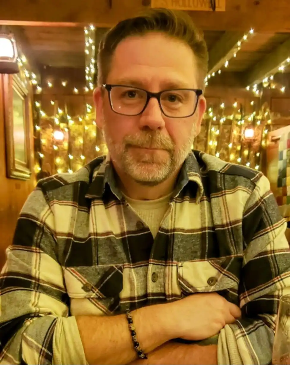 A middle-aged man with glasses and a beard, wearing a plaid shirt, sits with arms crossed in a warmly lit room decorated…
