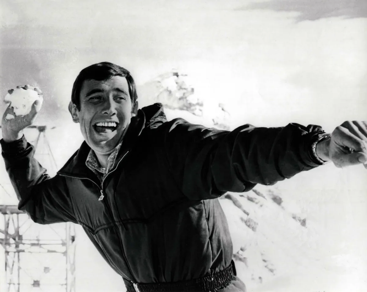 A black and white photo of a man in a winter jacket, smiling broadly while holding a snowball in one hand and extending the…