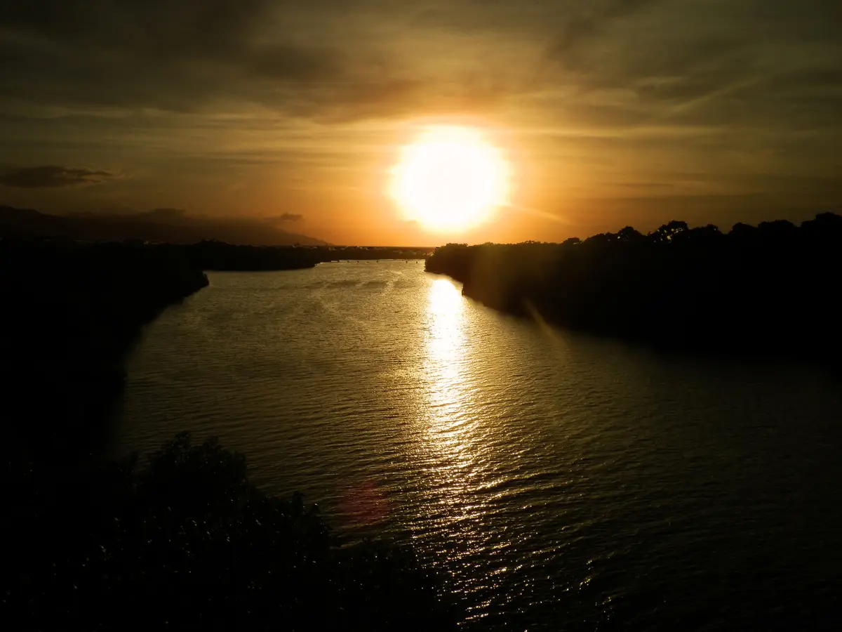 A serene sunset over a river with the sun casting a golden glow on the water, framed by silhouetted trees and distant hills…