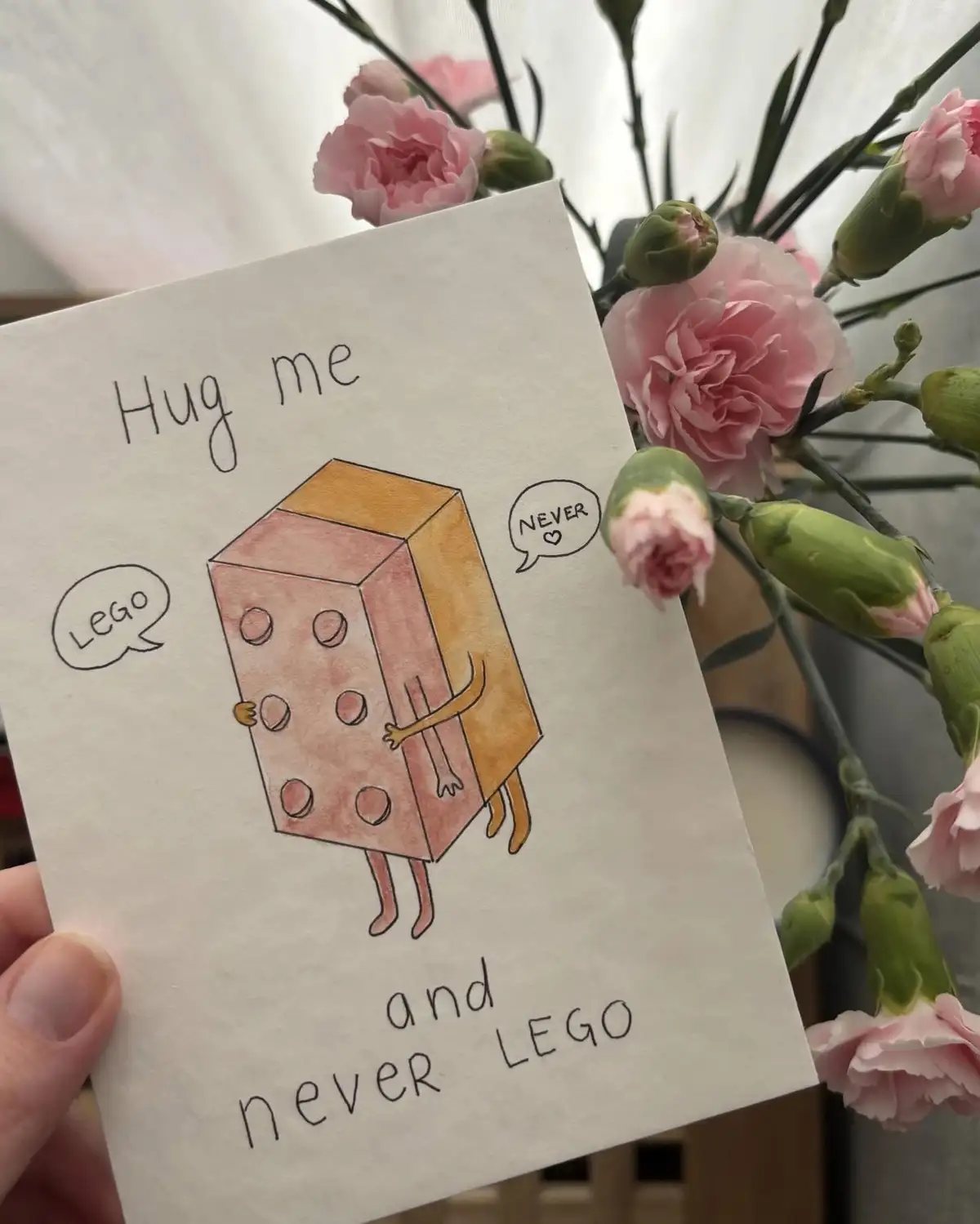 A hand holds a handmade card featuring a cartoon drawing of a pink and orange Lego brick with arms and legs. The brick has a…