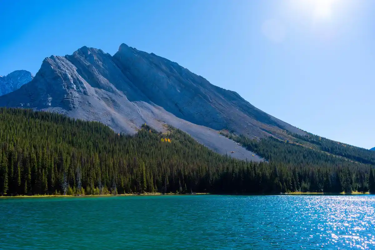 A scenic photo of a large mountain with a rocky peak and forested lower slopes, reflected in a turquoise lake under a bright…