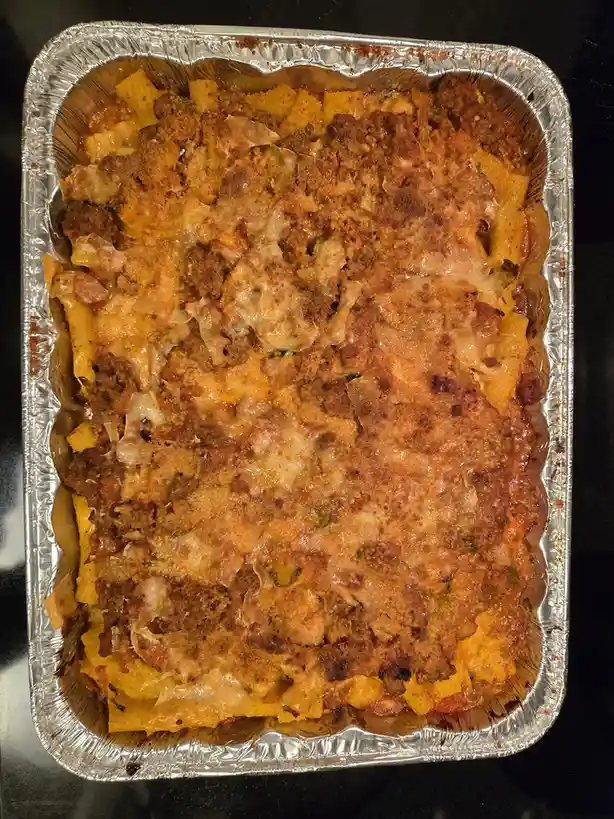 A top-down photo of a baked pasta dish in a rectangular aluminum foil pan, featuring golden-brown, bubbly cheese and meat…