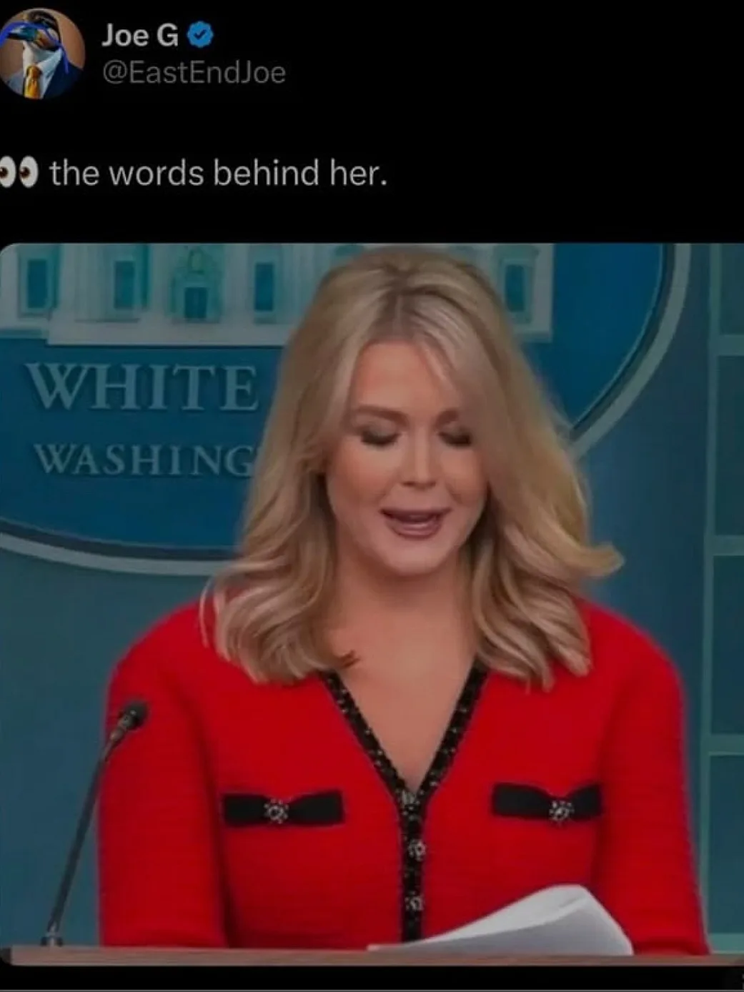 A woman with blonde hair wearing a red jacket stands at a podium in front of a blue backdrop that reads 'WHITE WASHING'. The…