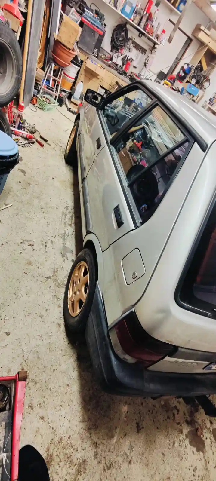 A silver compact car is parked inside a cluttered, messy home garage filled with tools, parts, and equipment. The car…