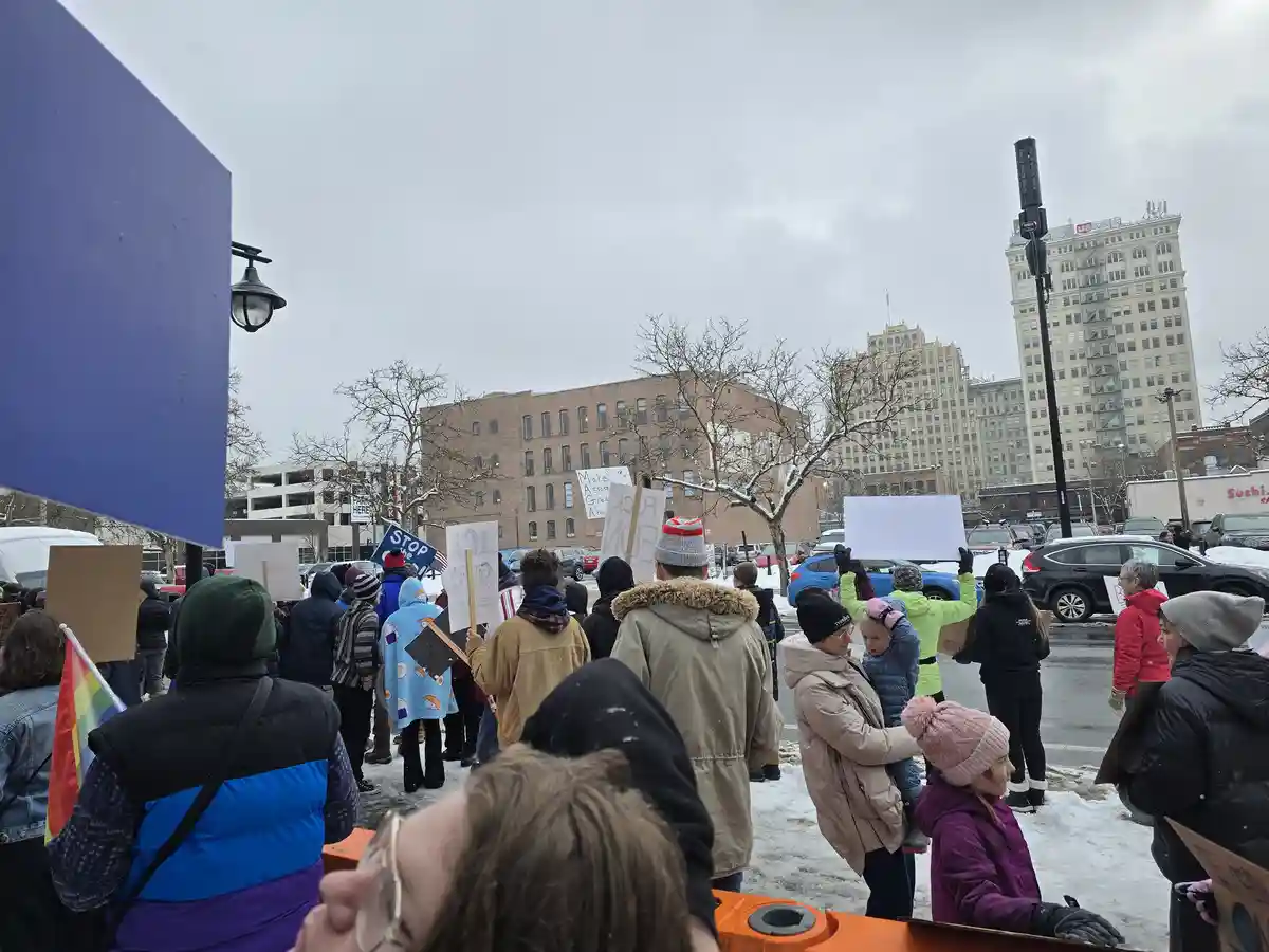 A snowy outdoor protest scene with many people holding signs, some visible signs include 'STOP ICE' and 'Make A ssosss Great…