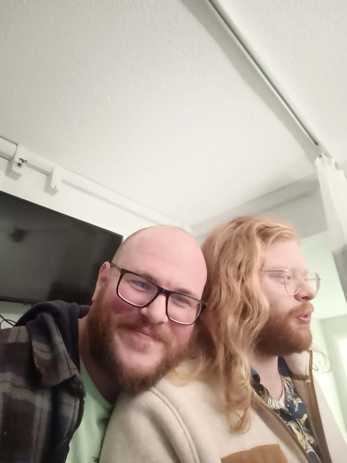 A selfie shows two men indoors, one bald with a beard and black-rimmed glasses smiling at the camera, while the other with…