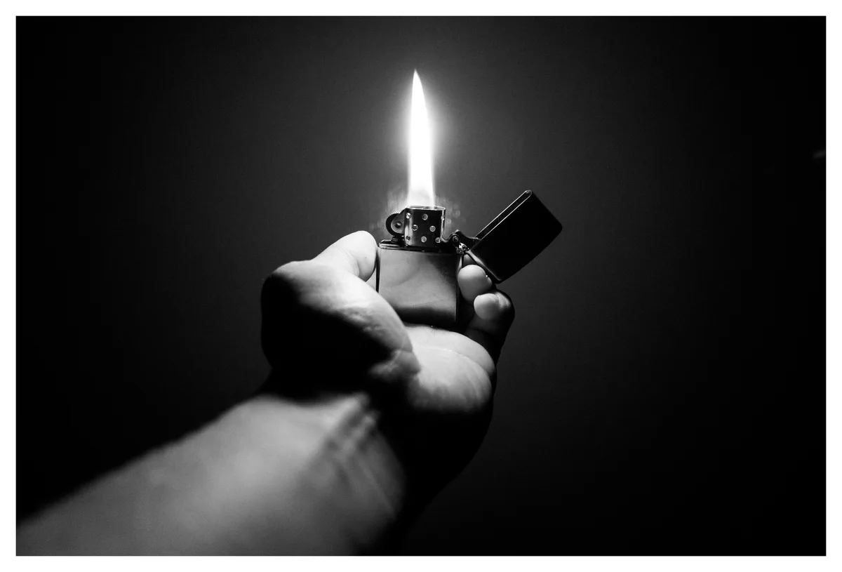 A black and white photo shows a hand holding a lit lighter against a dark background, with the flame casting light on the…