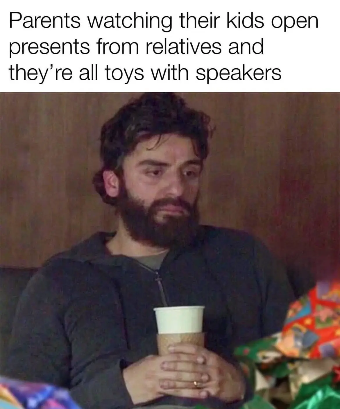 A man with a beard and dark hair, wearing a dark hoodie, sits holding a paper cup with both hands. He has a weary, resigned…