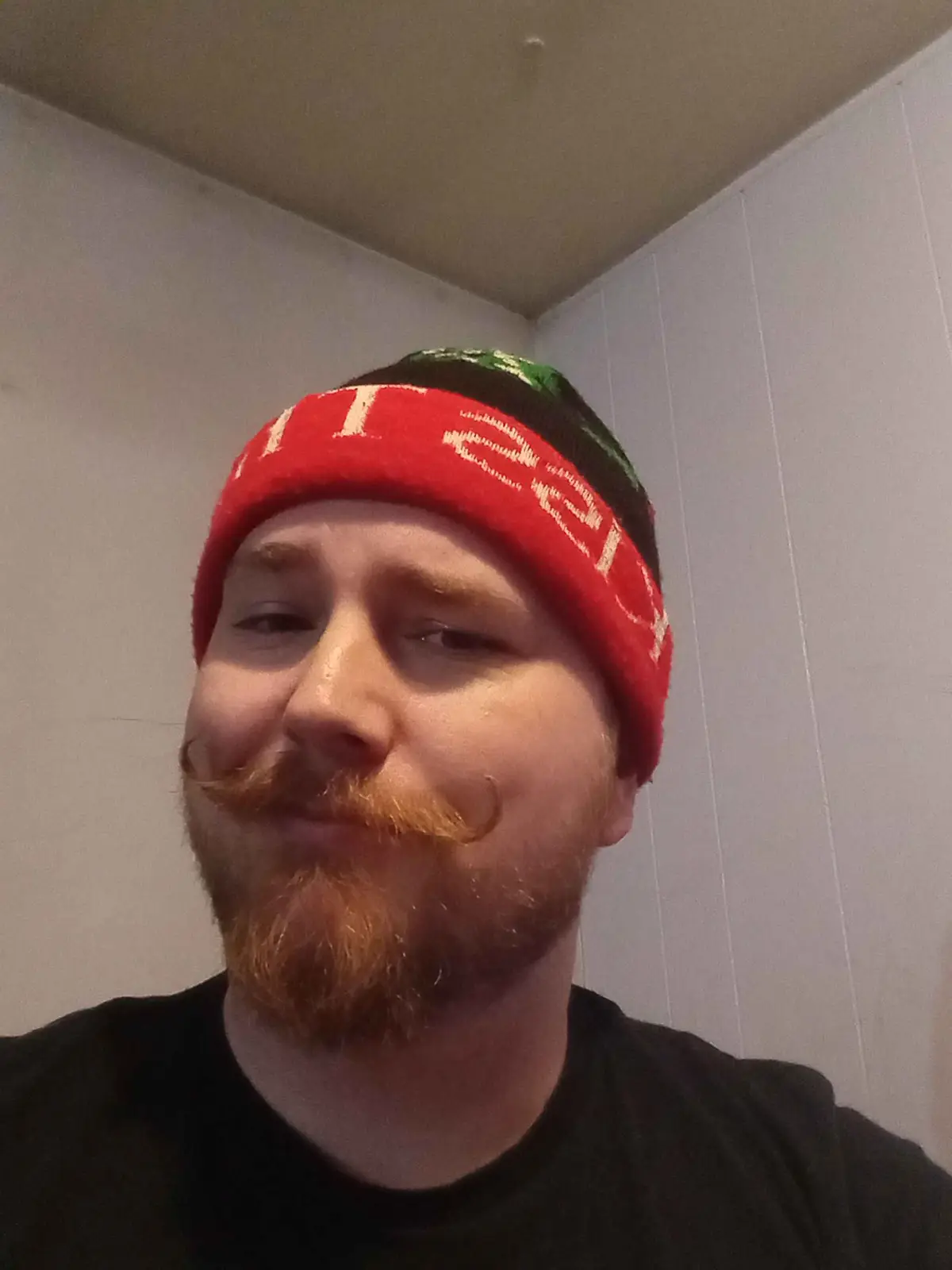 A man with a reddish beard and a distinctive handlebar mustache wears a red, black, and green knit beanie with white text…