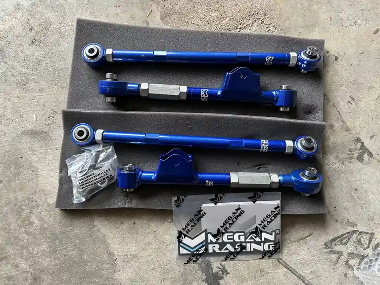 An overhead shot displays two sets of blue Megan Racing suspension control arms and tie rods neatly arranged on grey foam…