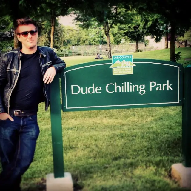 A man in sunglasses and a leather jacket leans casually against a green park sign that reads 'Dude Chilling Park' with a…
