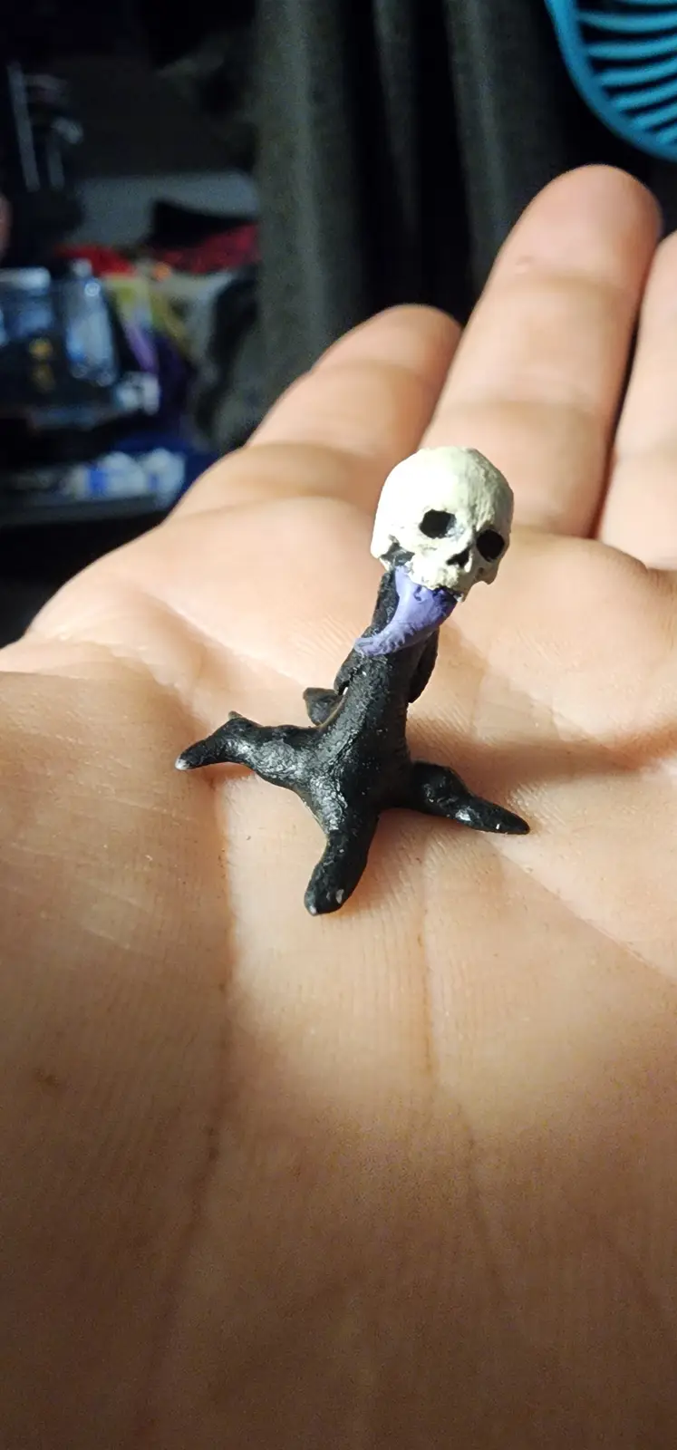 A small, handmade figurine of a skeletal bird with a skull head and a purple tongue is held in a person's open palm. The…