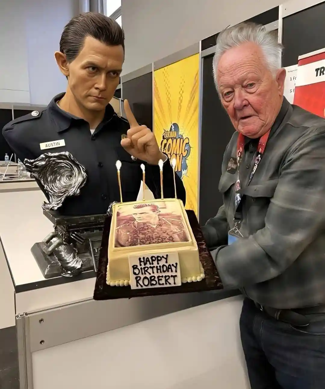 An elderly man stands beside a realistic wax figure of actor Robert Pattinson, who is pointing a finger at him. Between them…