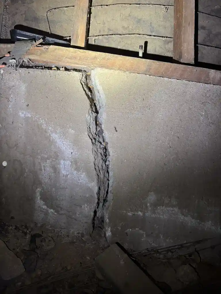 A close-up photo shows a large, jagged vertical crack running through a concrete basement wall, with exposed wood framing…