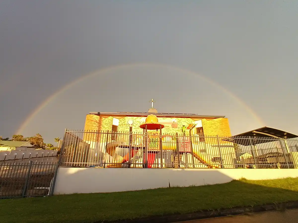 A brick church with a cross on its roof is framed by a vivid rainbow arching across the sky, with a colorful playground…